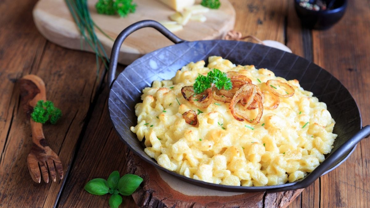 Käsespätzle nach klassischen Rezept in einer gusseiserenen Pfanne. Die steht auf einem rustikalen Untergrund aus Holz.