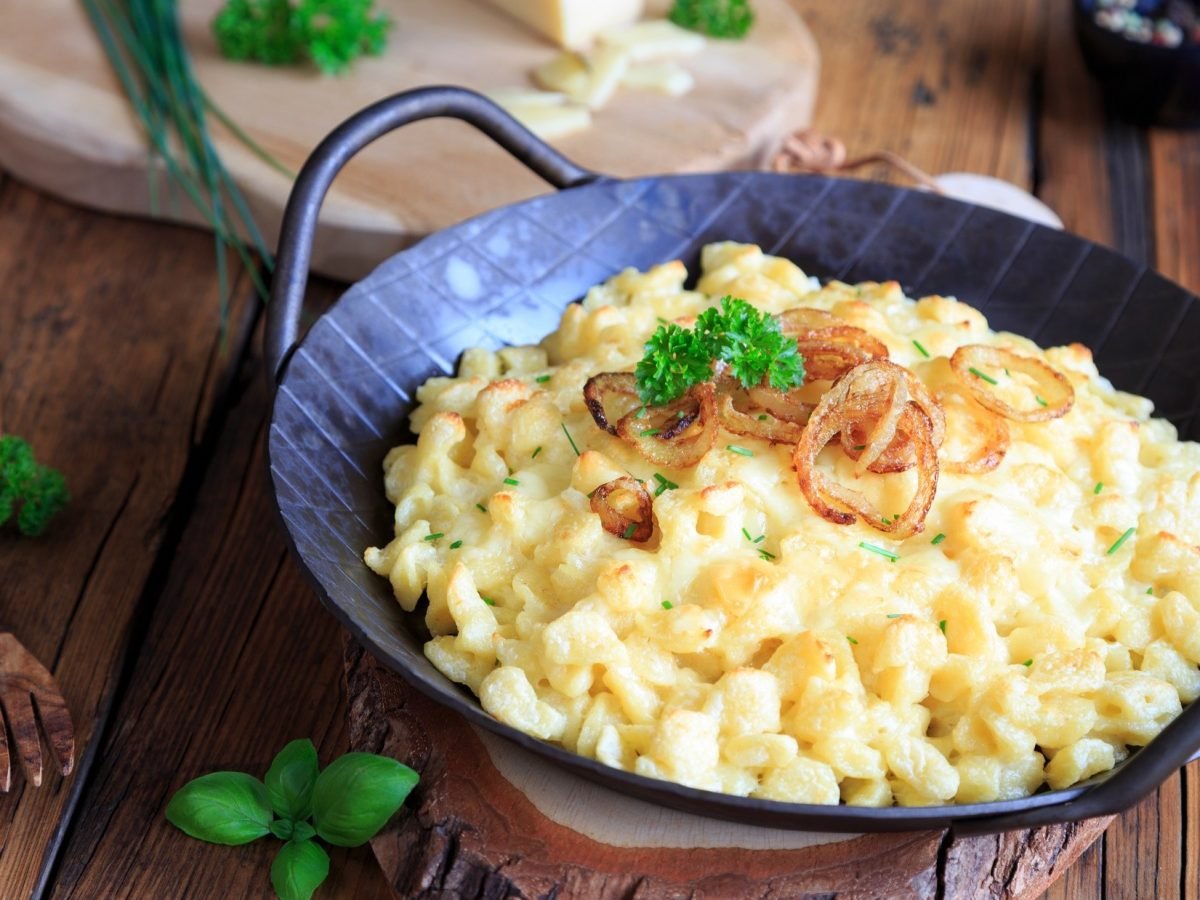 Käsespätzle nach klassischen Rezept in einer gusseiserenen Pfanne. Die steht auf einem rustikalen Untergrund aus Holz.