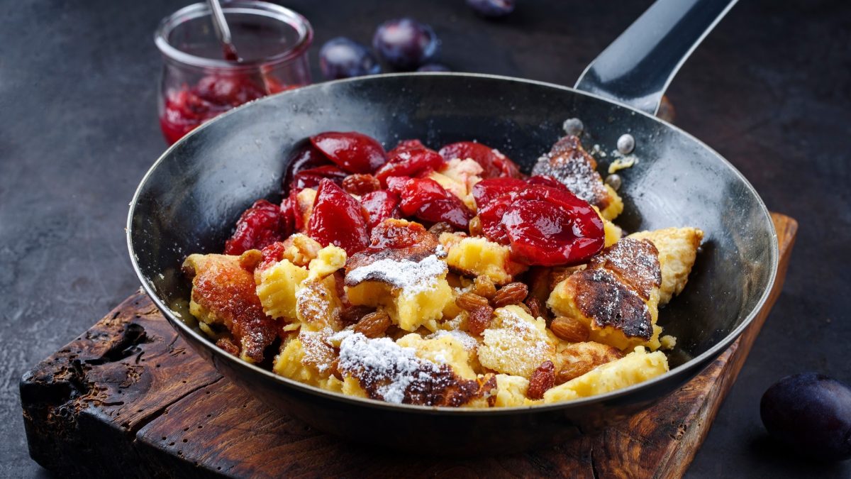 Draufsicht schräg: Ein Kaiserschmarrn mit Zwetschgenröster in einer schwarzes gusseisernen Pfanne auf einem dunklen und massivem Holzbrett. / Kaiserschmarrn – So gelingt er immer