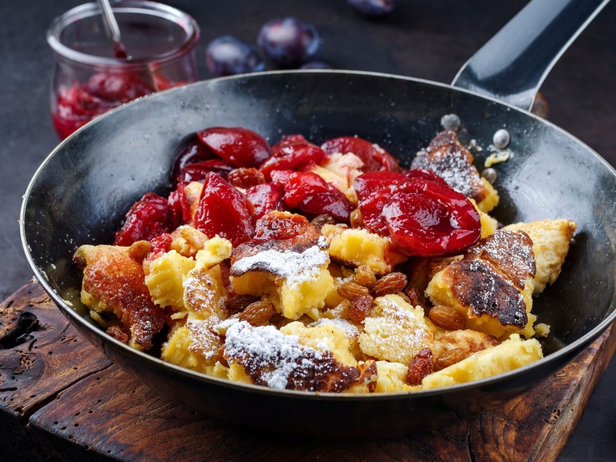 Draufsicht schräg: Ein Kaiserschmarrn mit Zwetschgenröster in einer schwarzes gusseisernen Pfanne auf einem dunklen und massivem Holzbrett. / Kaiserschmarrn – So gelingt er immer