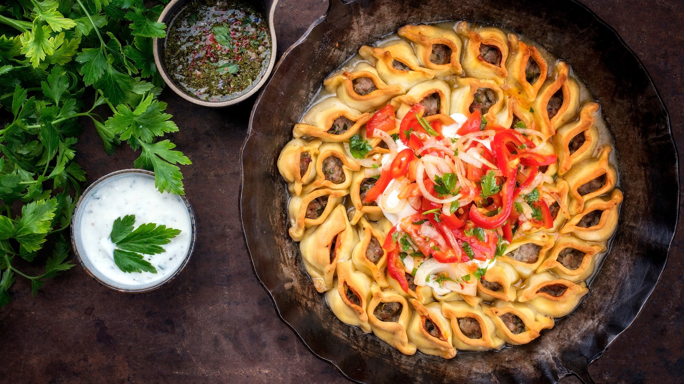 Manti mit Hackfleisch in einer ofenfesten gusseisernen runden Form. Die steht auf einem dunklen Untergrund. Drum herum ist eine Sauce aus Joghurt, eine Schale mit einer Sauce und frische Petersilie.