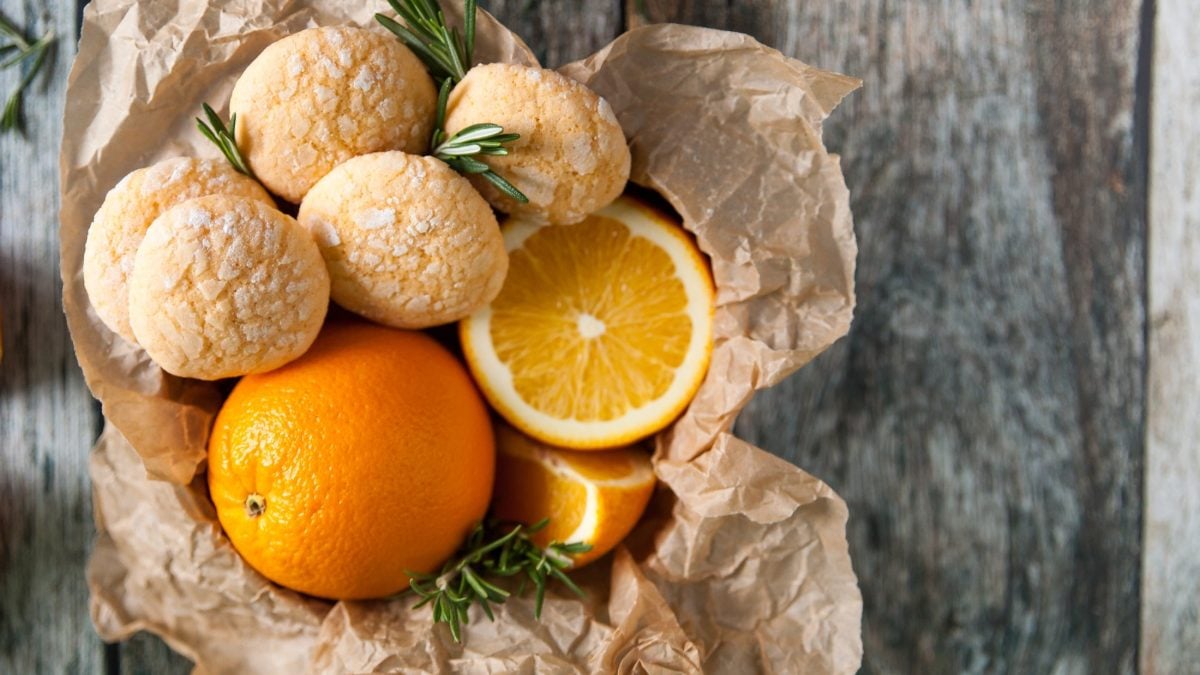 Orangenkekse und frische Orangen in einem Korb. Der steht auf einem hellen, melierten Untergrund.