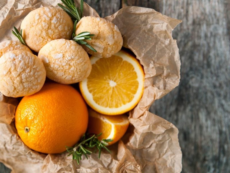 Orangenkekse und frische Orangen in einem Korb. Der steht auf einem hellen, melierten Untergrund.