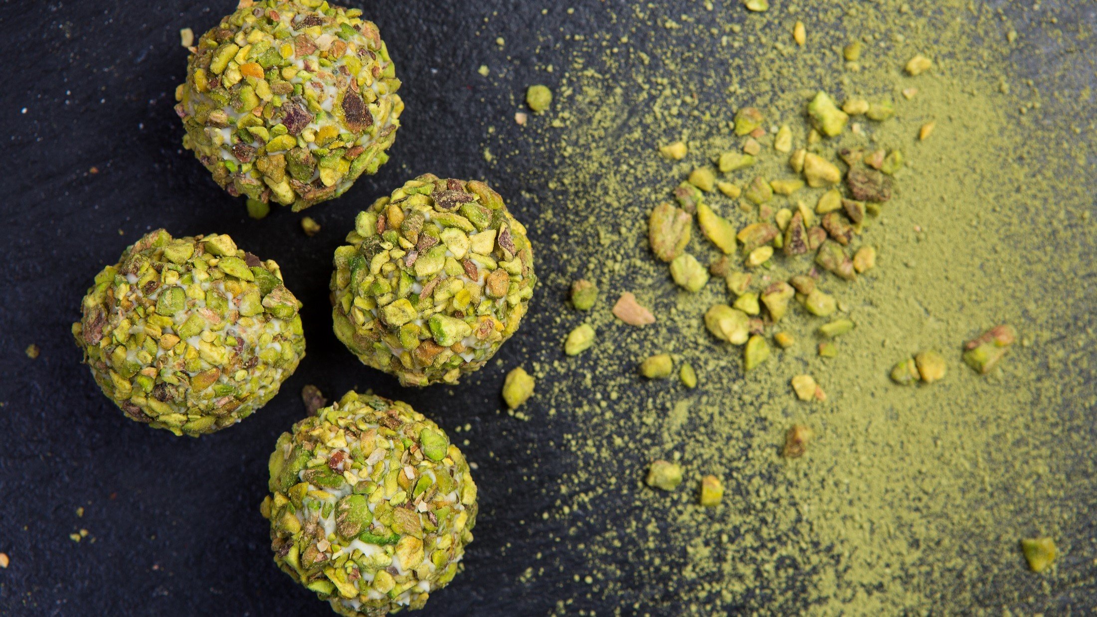 Pistazien-Energy-Balls auf einem schwarzen Untergrund. Drum herum sind klein gehackte Pistazien sowie ein grünes Pulver.