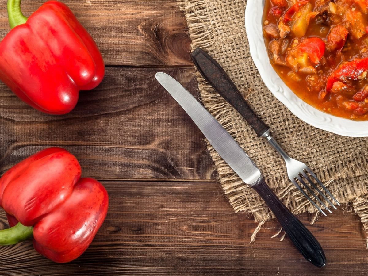 Draufsicht Ein weißer Teller mit einer selbst gemachten Reitersuppe Daneben liegen zwei rote Paprika und ein Besteck.