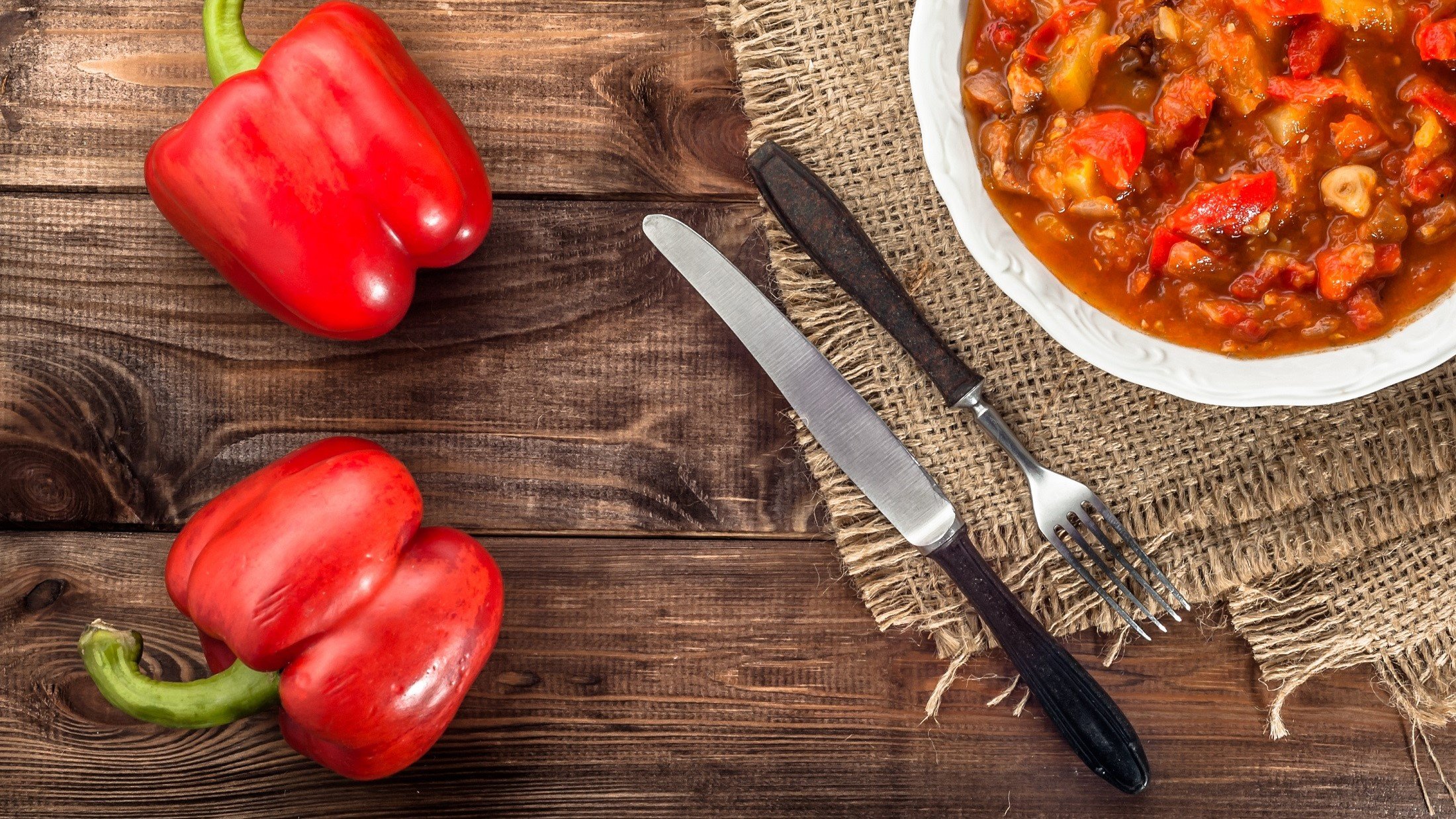 Draufsicht Ein weißer Teller mit einer selbst gemachten Reitersuppe Daneben liegen zwei rote Paprika und ein Besteck.