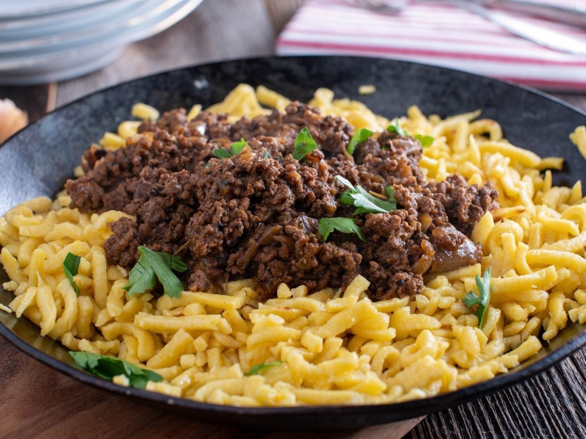 Draufsicht, schräg: eine rustikale Spätzlepfanne mit Hackfleisch auf einem Untergrund aus Holz. Im Hintergrund sind Küchentücher und weitere Teller.