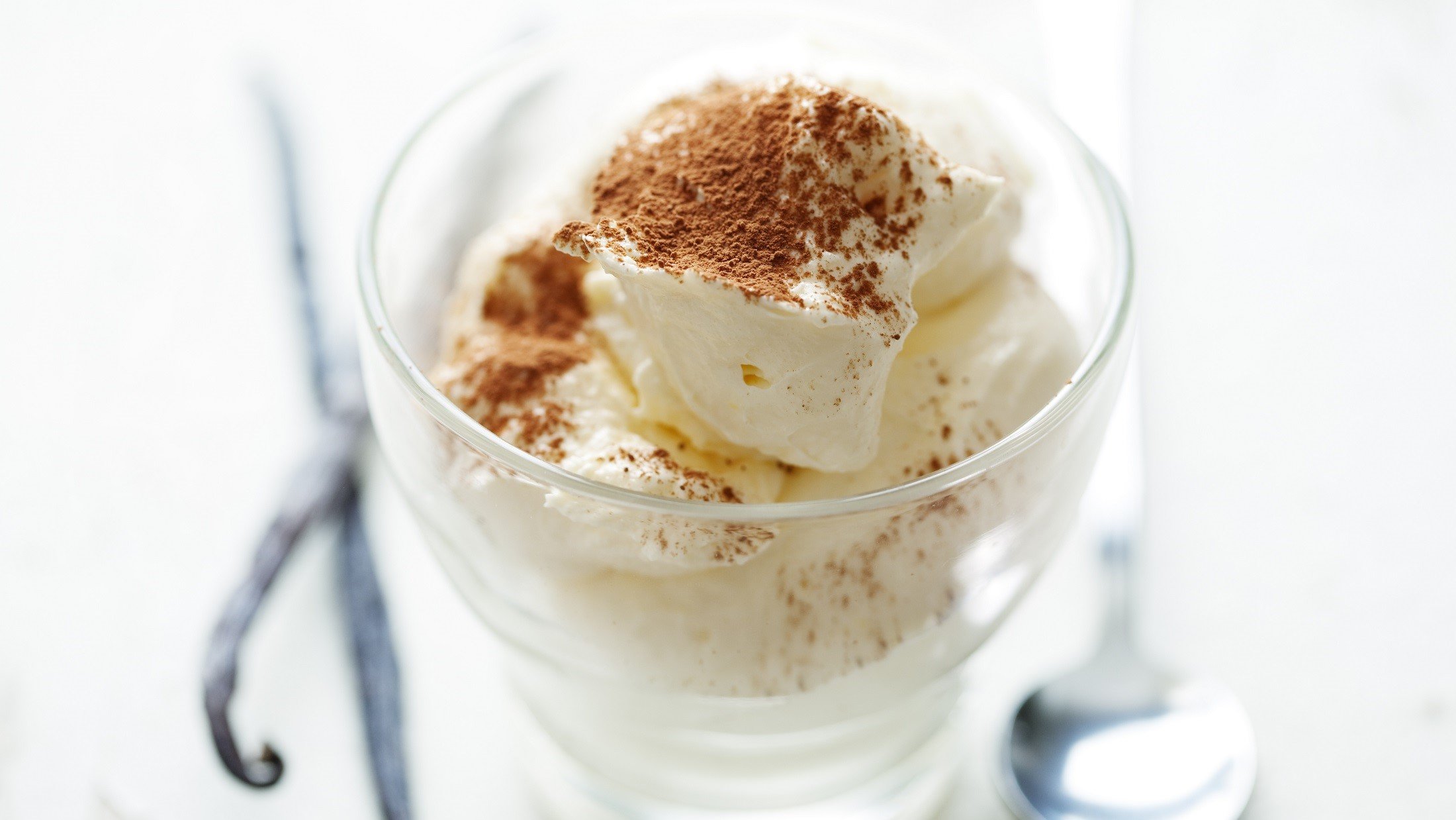 Vanille-Mousse mit Kakaopulver bestreut in einem Dessertglas. Auf dem weißen Untergrund liegen drei schmale Vanilleschoten. Draufsicht, schräg.