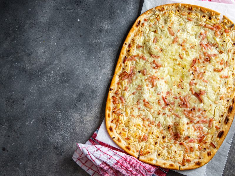 Knuspriger Flammkuchenteig mit Belag vor grauem Untergrund von oben fotografiert.