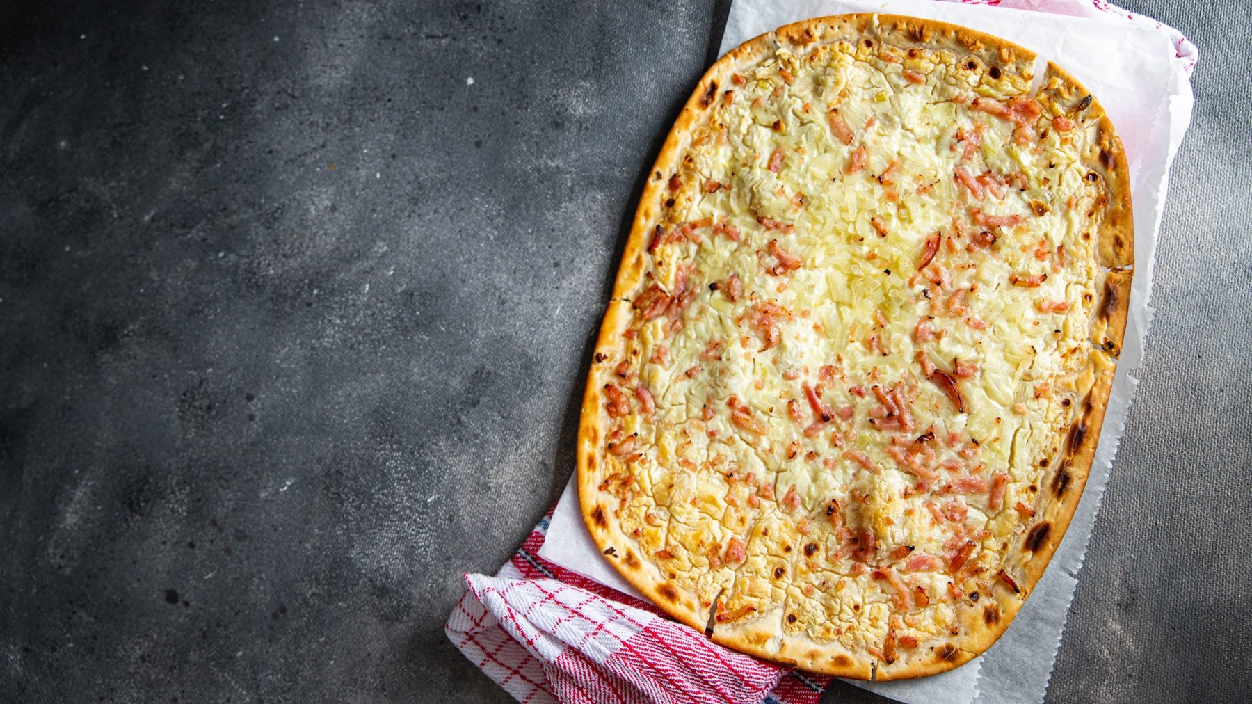 Knuspriger Flammkuchenteig mit Belag vor grauem Untergrund von oben fotografiert.