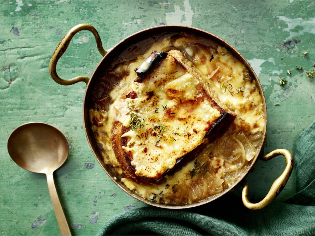 Draufsicht: Französische Zwiebelsuppe im Topf, daneben ein Löffel auf grünem Holzuntergrund.