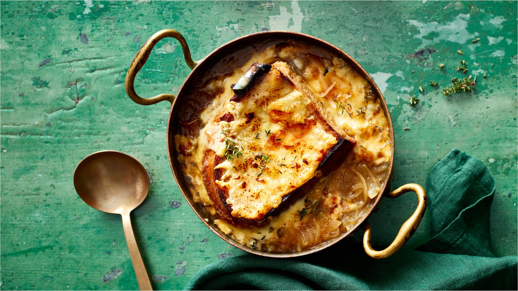 Draufsicht: Französische Zwiebelsuppe im Topf, daneben ein Löffel auf grünem Holzuntergrund.