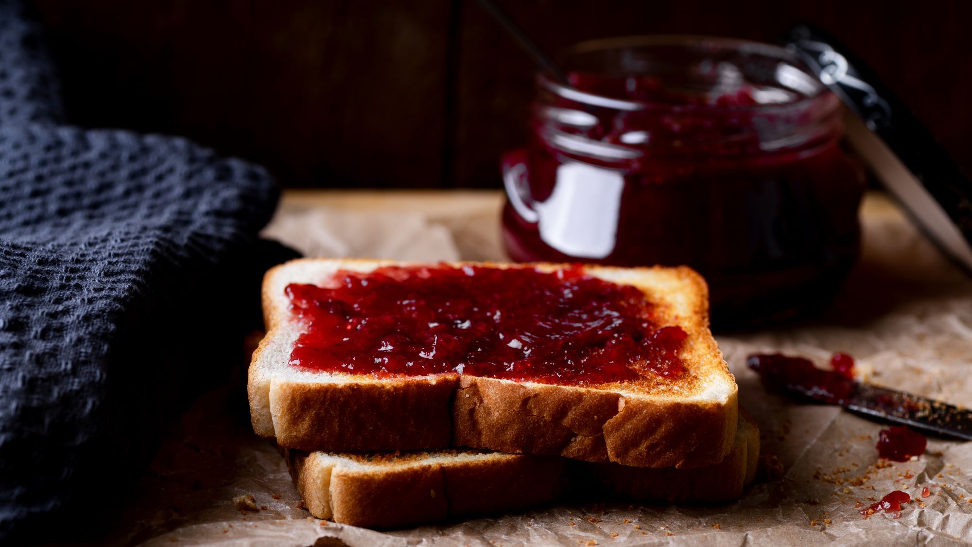 Zwei goldbraune Toastscheiben mit leuchtender Glühweinmarmelade, daneben ein Glas Konfitüre und ein Messer auf braunem Papier.