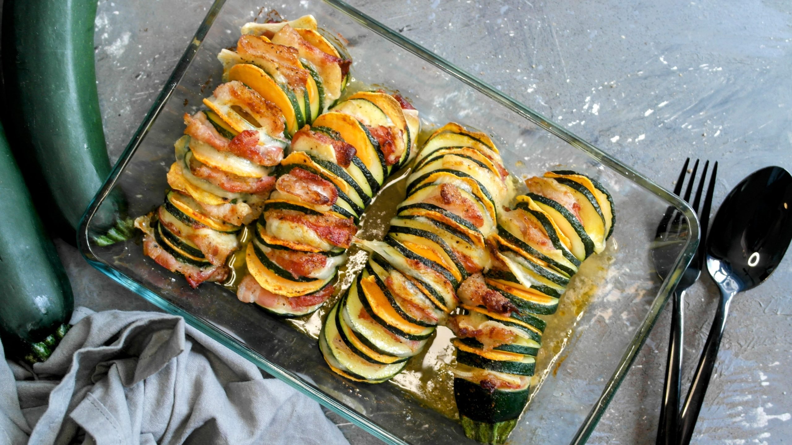 Eine Glasauflaufform mit der überbackenen Hasselback-Zucchini auf grauem Steinuntergrund. Alles direkt von oben fotografiert.