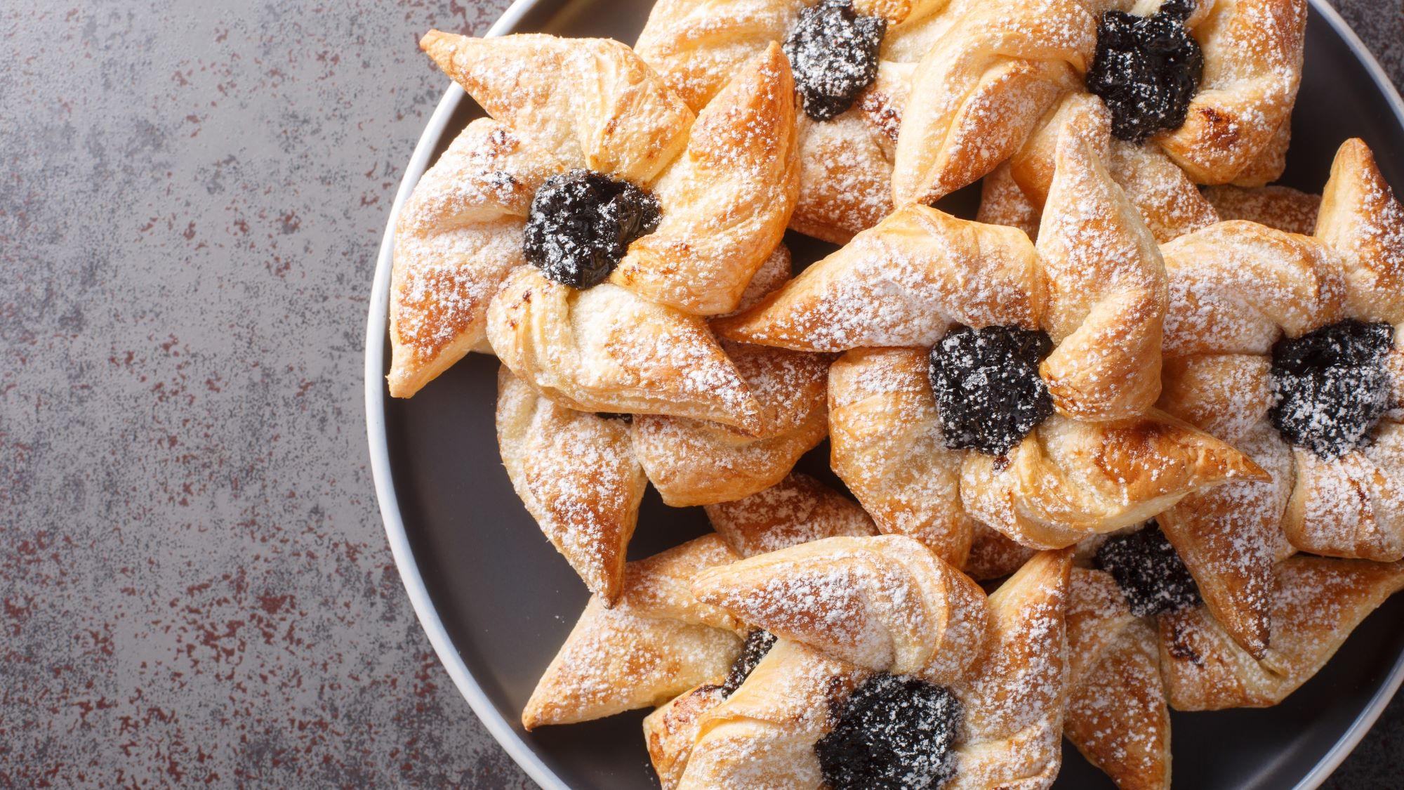 Stapel aus sternförmigem Blätterteiggebäck namens Joulutorttu mit Pflaumenmusfüllung und Puderzucker bestäubt auf grauem Teller.