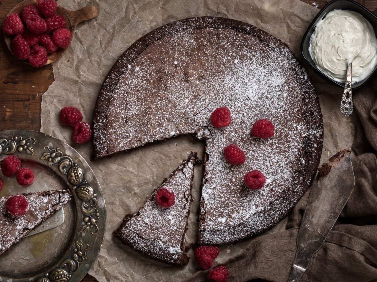 Schokoladenkuchen Kladdkaka mit Puderzucker bestäubt und frischen Himbeeren garniert, ein Stück auf antikem Teller, daneben Sahnecreme.