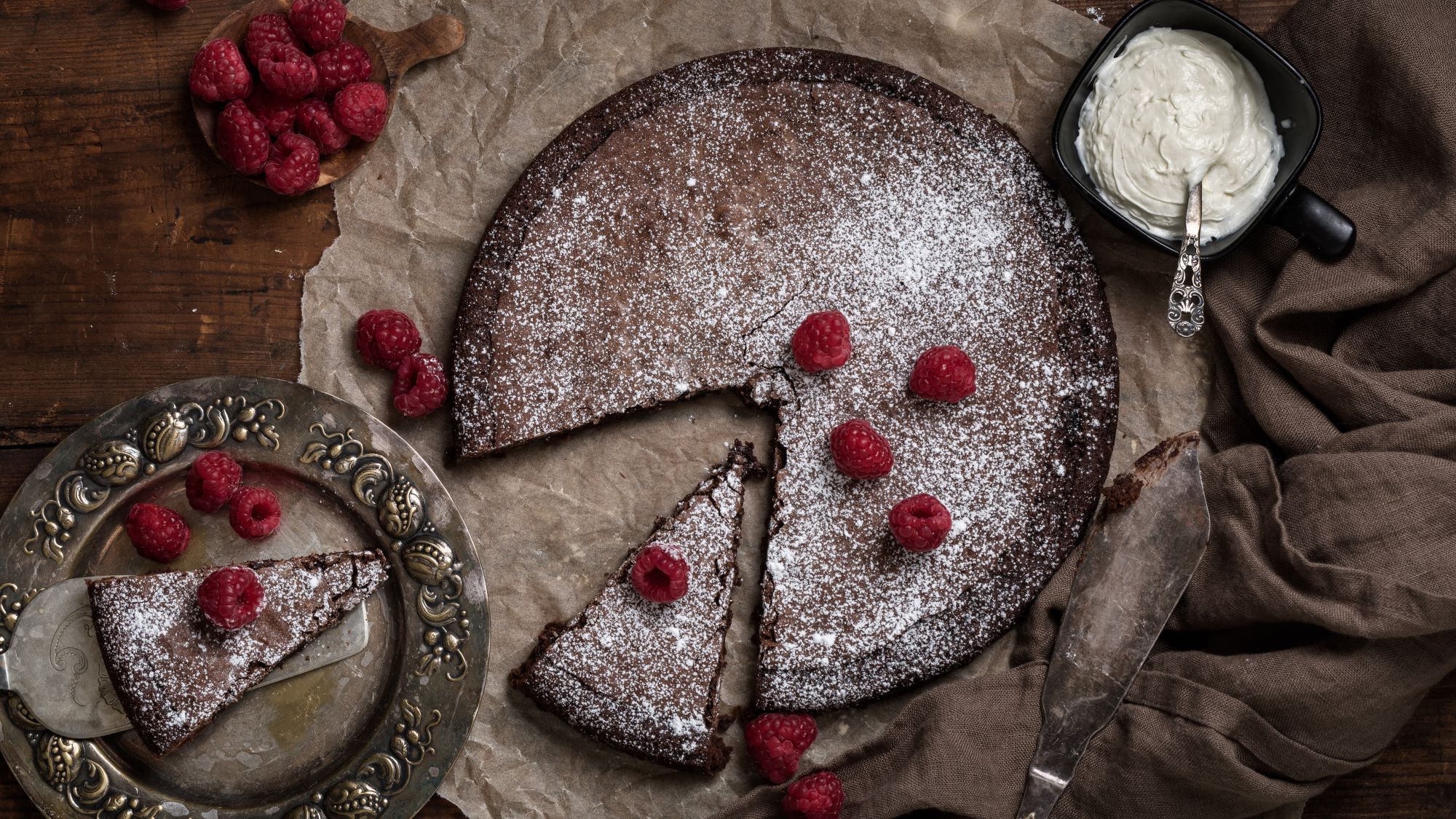 Schokoladenkuchen Kladdkaka mit Puderzucker bestäubt und frischen Himbeeren garniert, ein Stück auf antikem Teller, daneben Sahnecreme.