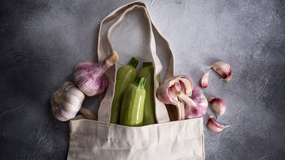 Frische grüne Zucchini und lilafarbene Knoblauchknollen ragen aus einer beigen Tasche, daneben verstreute Knoblauchzehen auf Steinboden.