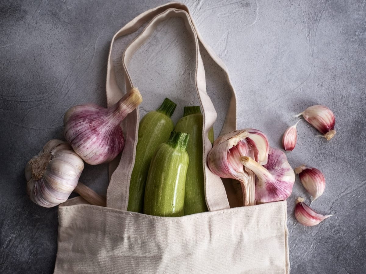 Frische grüne Zucchini und lilafarbene Knoblauchknollen ragen aus einer beigen Tasche, daneben verstreute Knoblauchzehen auf Steinboden.