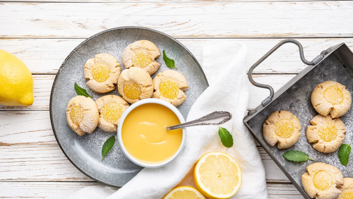 Die Lemon Curd Plätzchen auf einem grauen Teller und weißem Holztisch. Drumherum Zitronen und Blätter der Pflanze zusammen mit den Grundzutaten als Deko. Alles direkt von oben fotografiert.