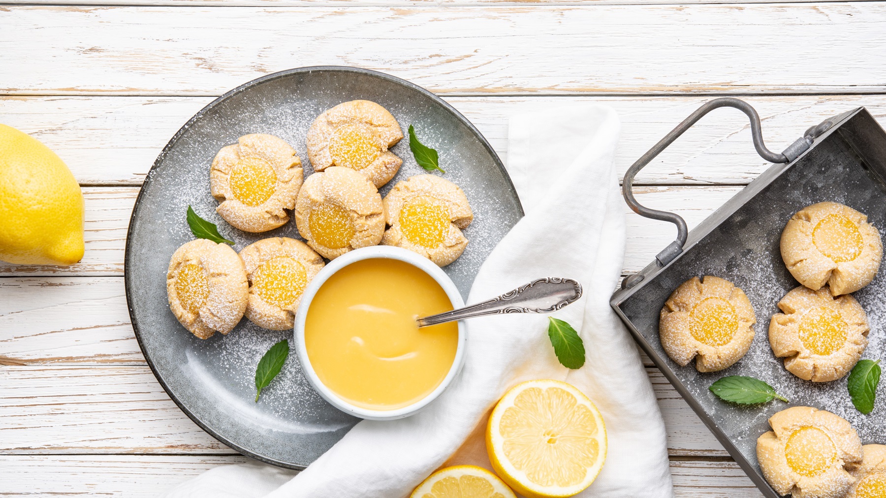 Die Lemon Curd Plätzchen auf einem grauen Teller und weißem Holztisch. Drumherum Zitronen und Blätter der Pflanze zusammen mit den Grundzutaten als Deko. Alles direkt von oben fotografiert.