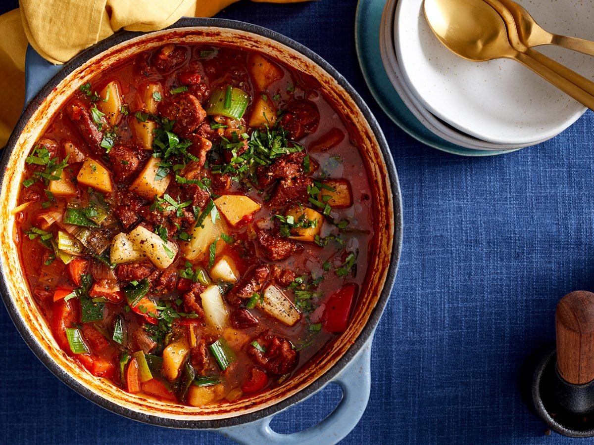 Draufsicht: Ofen-Gulaschsuppe in einem Topf, daneben Schüsseln und Löffel auf dunkelblauem Untergrund.