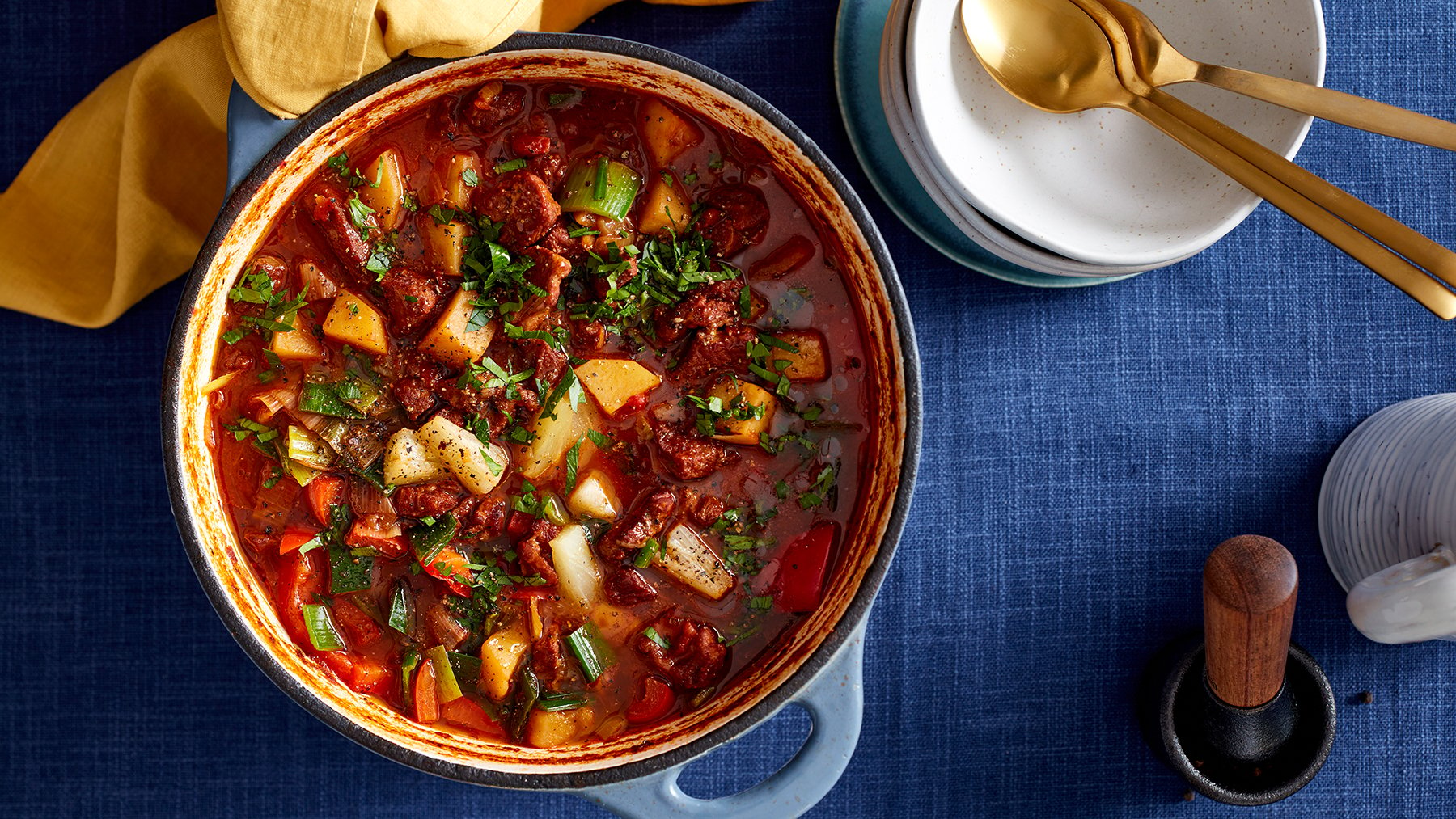 Draufsicht: Ofen-Gulaschsuppe in einem Topf, daneben Schüsseln und Löffel auf dunkelblauem Untergrund.