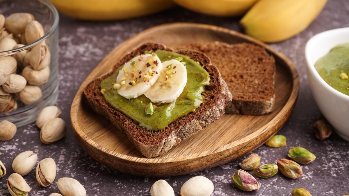 Eine Scheibe Vollkornbrot die mit Pistazienbutter und Bananenscheiben belegt ist, liegt auf einem rustikalen Frühstücksteller. Auf der Holzoberfläche liegen frische Pistazien und Bananen.