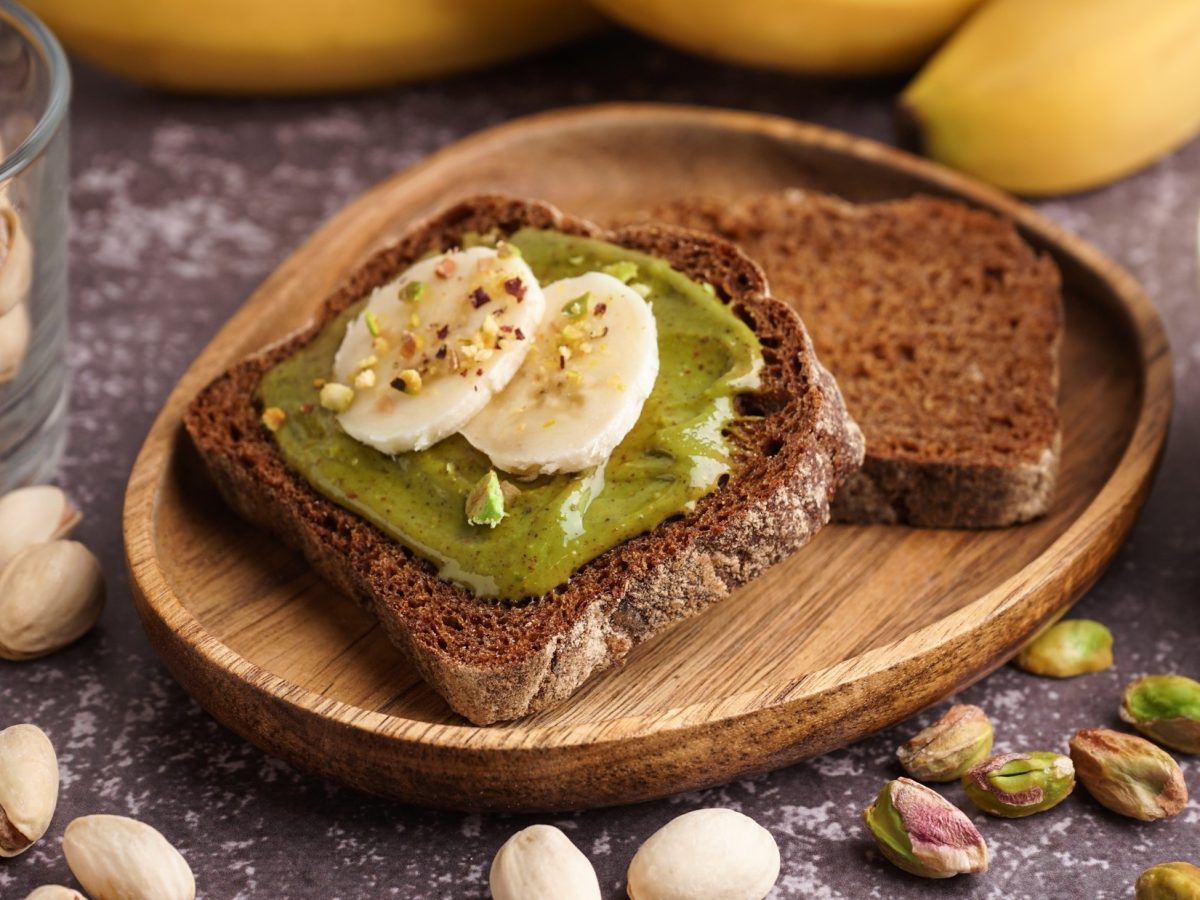 Eine Scheibe Vollkornbrot die mit Pistazienbutter und Bananenscheiben belegt ist, liegt auf einem rustikalen Frühstücksteller. Auf der Holzoberfläche liegen frische Pistazien und Bananen.