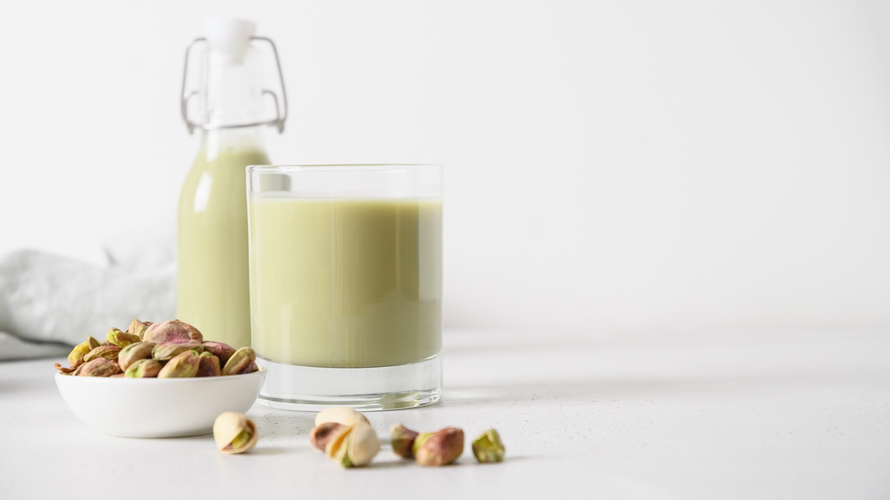 Ein Glas und eine Flasche mit Pistazienmilch vor einem weißen Hintergrund, im Vordergrund frische Pistazien.