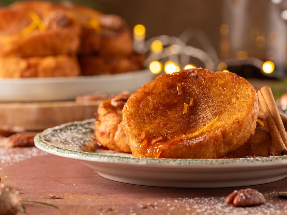 Rabanadas, goldbraun gebratene Brotscheiben, die mit Zimt, Zucker und einem Hauch von Orangenzesten verziert sind. Im Hintergrund sieht man weitere Portionen und weihnachtliche Dekorationen.