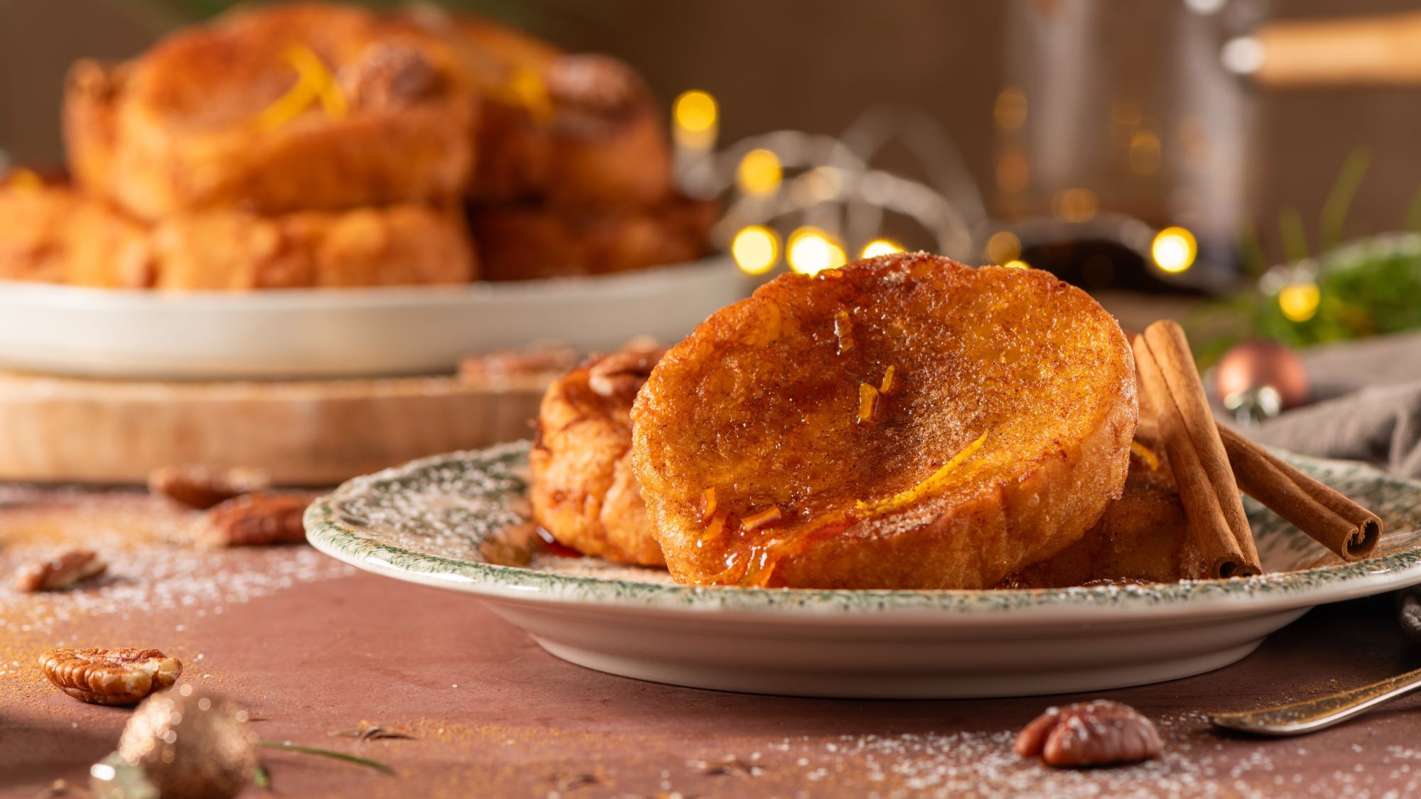 Rabanadas, goldbraun gebratene Brotscheiben, die mit Zimt, Zucker und einem Hauch von Orangenzesten verziert sind. Im Hintergrund sieht man weitere Portionen und weihnachtliche Dekorationen.