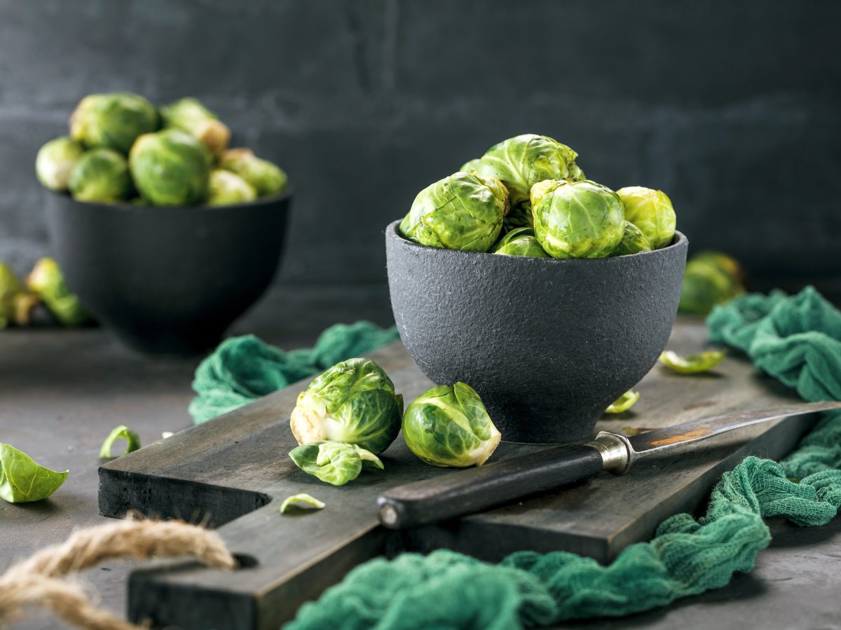 Rosenkohl vor dem Putzen in zwei grauen Schalen, die auf einem Brett stehen. Daneben ein dunkelgrünes Tuch.