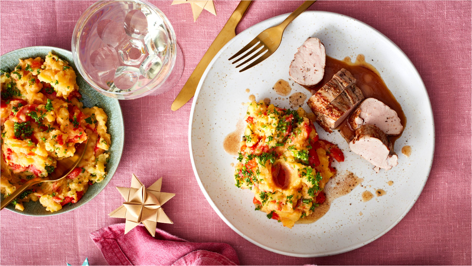 Draufsicht: Schweinefilet mit Paprika-Kartoffelpüree auf einem Teller, daneben ein Teller mit mehr Püree und ein Glas. Alles auf rosa Untergrund.