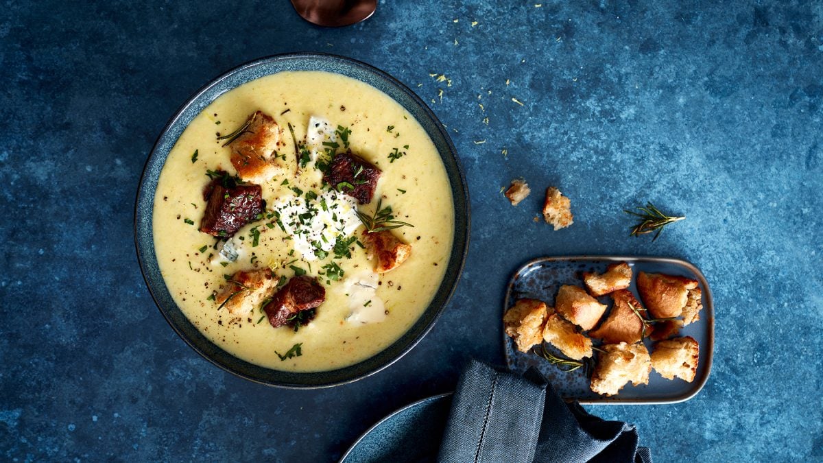 Zitronige Kartoffelsuppe mit Gorgonzola-Steak-Finish in einer dunkelblauen Schüssel auf dunkelblauem Hintergrund. Rechts ein kleines Schüsselchen mit Croûtons.