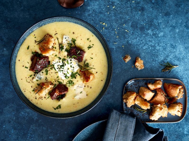 Zitronige Kartoffelsuppe mit Gorgonzola-Steak-Finish in einer dunkelblauen Schüssel auf dunkelblauem Hintergrund. Rechts ein kleines Schüsselchen mit Croûtons.