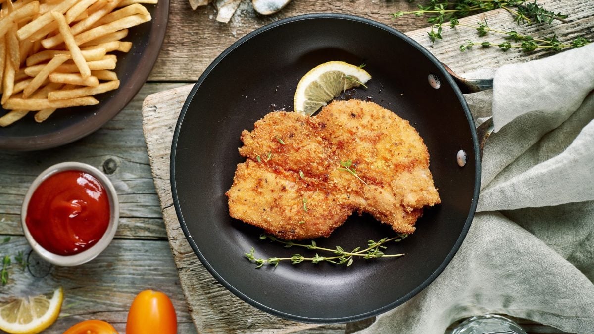 Thymian-Schnitzel auf schwarzem Teller. Daneben Zitrone und frischer Thymian, im Szenario Pommes und Ketchup.