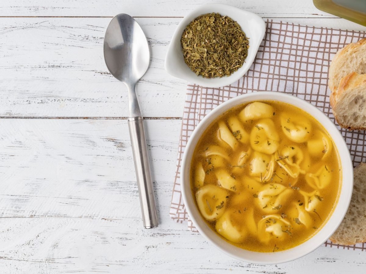 Tortellinisuppe in heller Brühe, serviert mit getrockneten Kräutern, frischem Brot und einem Löffel auf rustikalem Holztisch.