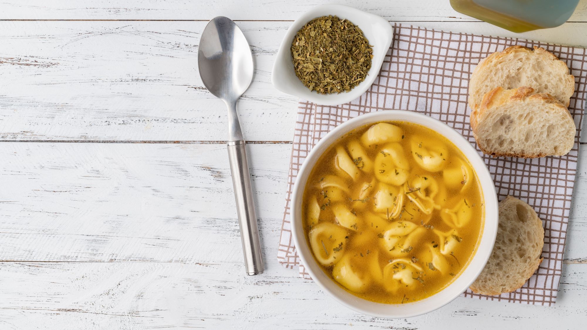 Tortellinisuppe in heller Brühe, serviert mit getrockneten Kräutern, frischem Brot und einem Löffel auf rustikalem Holztisch.