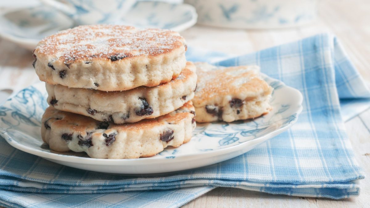 Auf dem Bild sind gestapelte Welsh Cookies zu sehen – flache, runde Kekse mit Rosinen, die leicht mit Zucker bestreut sind. Sie liegen auf einem Teller mit blauen Mustern, der auf einem blau-weiß karierten Tuch arrangiert ist.