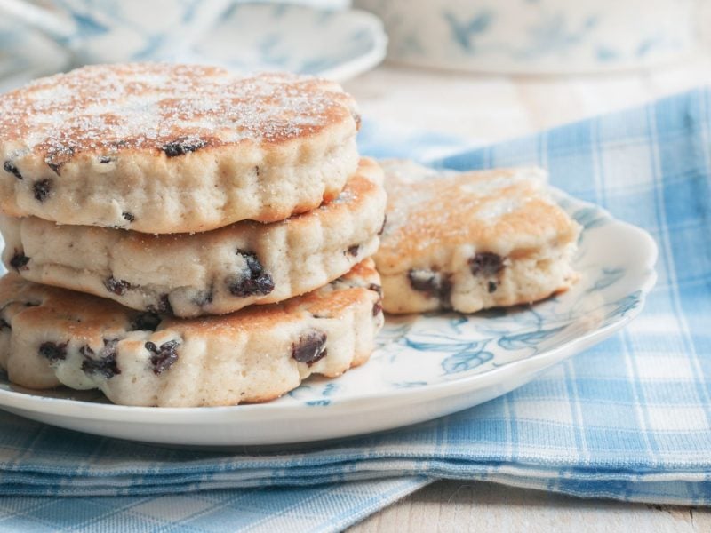 Auf dem Bild sind gestapelte Welsh Cookies zu sehen – flache, runde Kekse mit Rosinen, die leicht mit Zucker bestreut sind. Sie liegen auf einem Teller mit blauen Mustern, der auf einem blau-weiß karierten Tuch arrangiert ist.