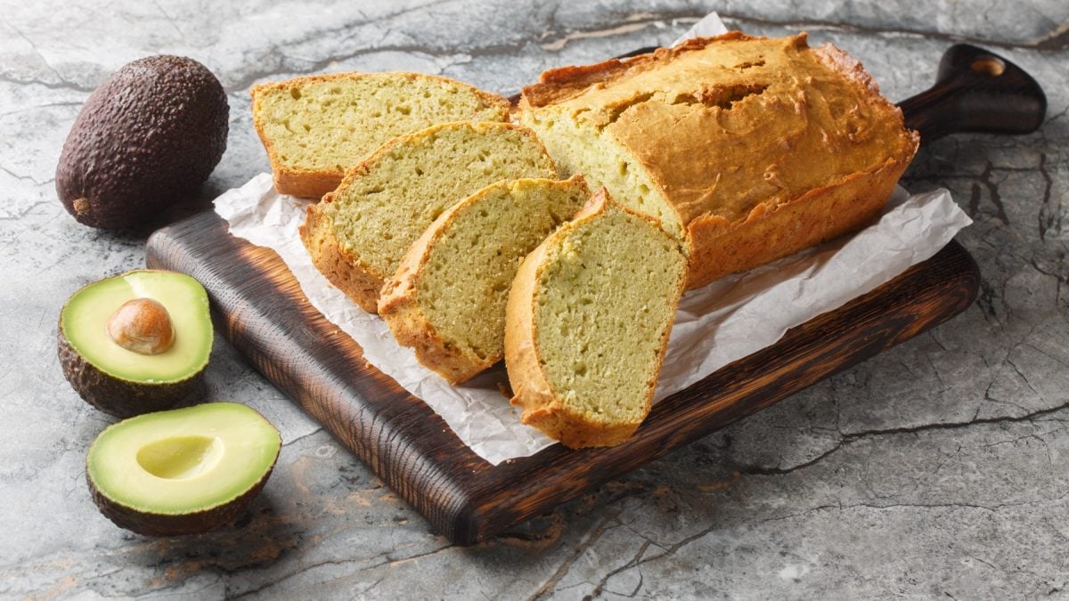 Angeschnittenes Avocadobrot auf einem Holzbrett, daneben eine halbierte Avocado und eine Ganze.