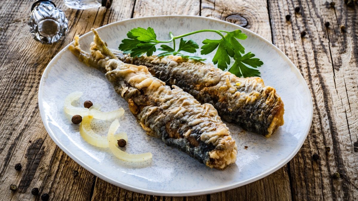 Draufsicht schräg: Ein weißer Teller mit zwei Bratheringen liegt auf einem rustikalen Untergrund aus Holz. neben den Heringsfilets liegen rohe Zwiebelringe und frische Petersilie.