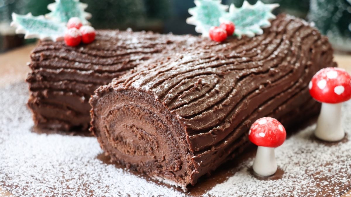 Bûche de Noel ohne Mehl auf Puderzucker, rechts davon Zuckerfiguren.