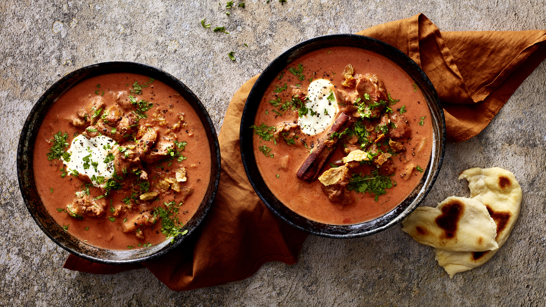 Zwei Schüsseln Butter-Chicken-Soup, daneben Stücke Fladenbrot auf grauem Untergrund.