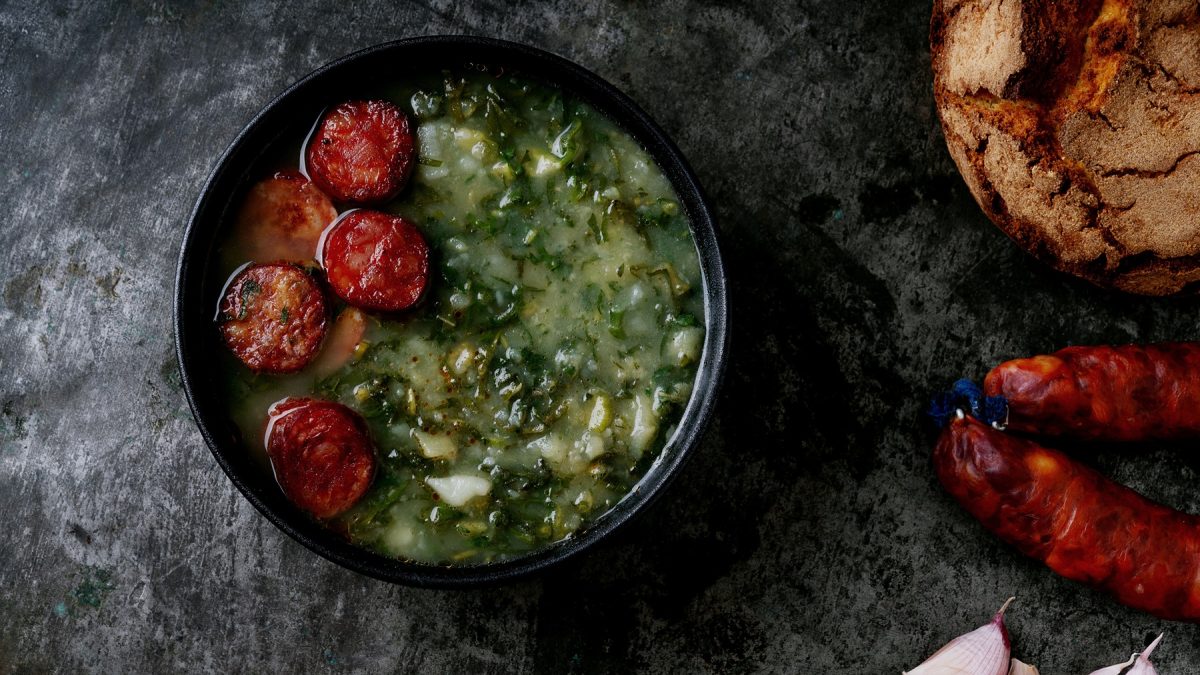 Eine schwarze Schale mit der Suppe auf einem grauen Steintisch, mit Knoblauchzehen, Chorizo und Sauerteigbrot im rechten Bildrand. Alles direkt von oben fotografiert.