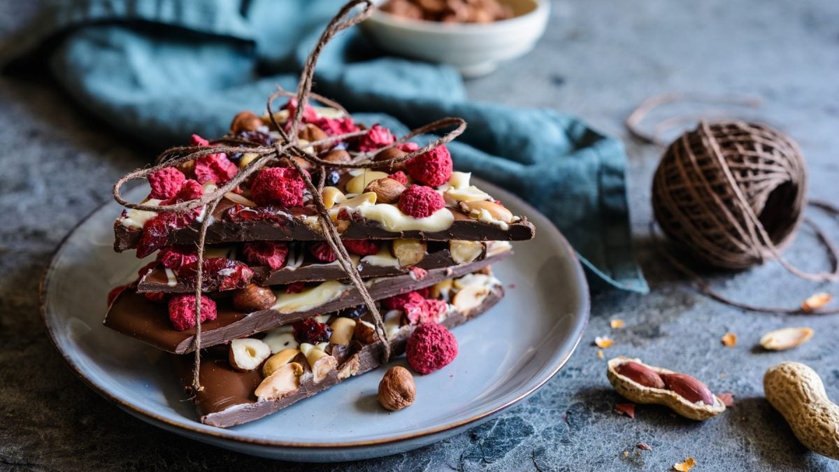 Frontalsicht auf mehrere Stücke Chocolate Bark mit Trockenfrüchten und Nüsse, die zu einem Paket geschnürt sind. Daneben eine Spule mit Paketband.