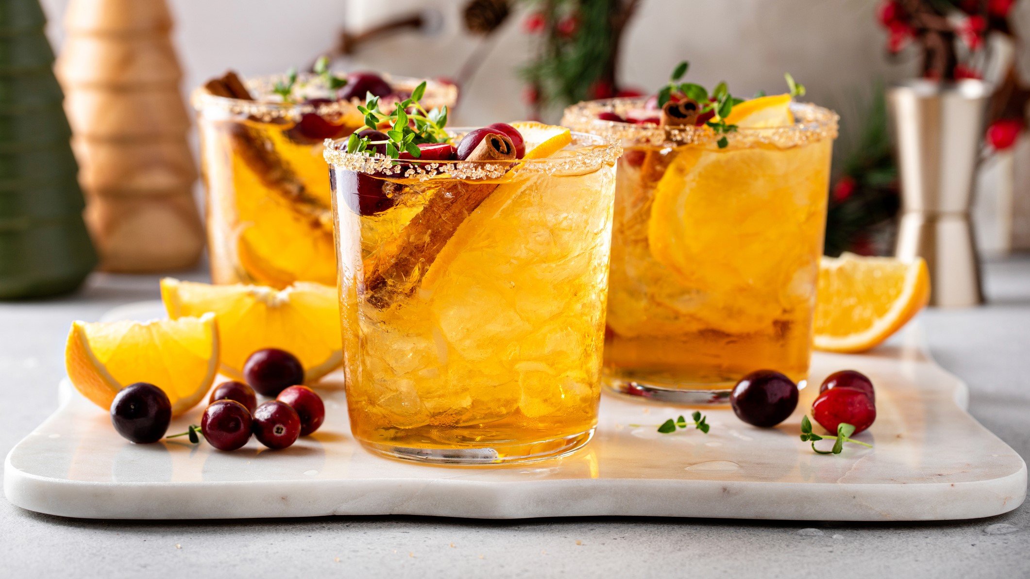 Drei Gläser mit Cranberry-Orangen-Mocktail auf einem Tablett mit frischen Orangenschnitzen und Cranberry daneben.