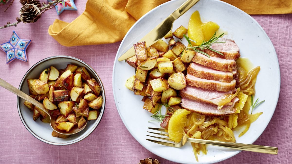 Draufsicht: Entenbrust mit Orangensauce und Bratkartoffeln auf einem Teller, daneben Schüssel mit mehr Bratkartoffeln, rosa Untergrund.