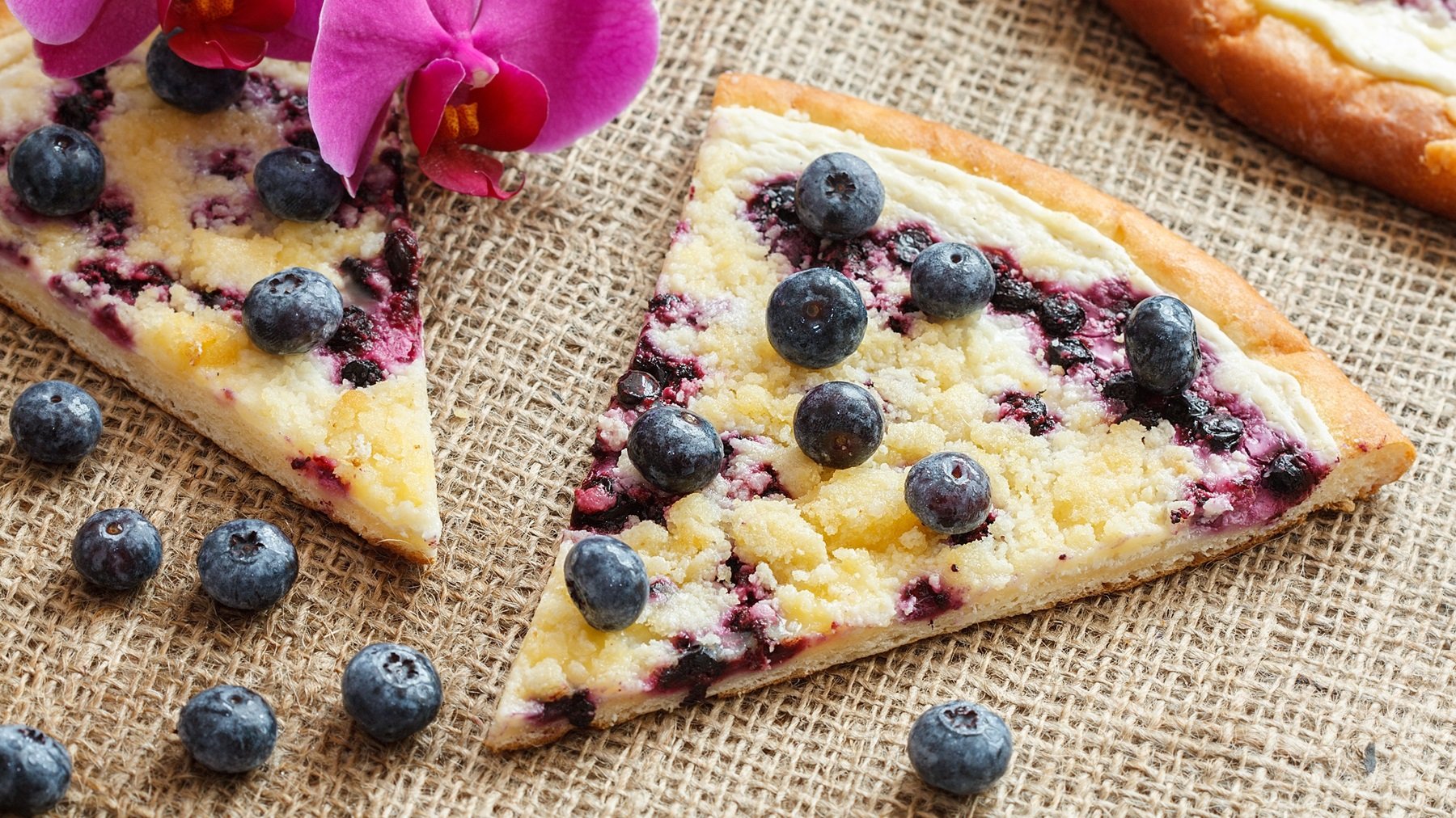 Stück Frgál mit Blaubeeren und Streusel auf einem Sisal-Untergrund, im Hintergrund eine pinke Orchidee.